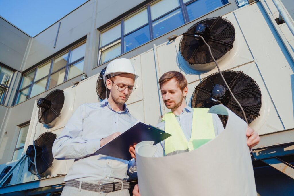 Two HVAC technicians reviewing installation plans in front of large outdoor cooling units, emphasizing eco-friendly HVAC solutions and system optimization.