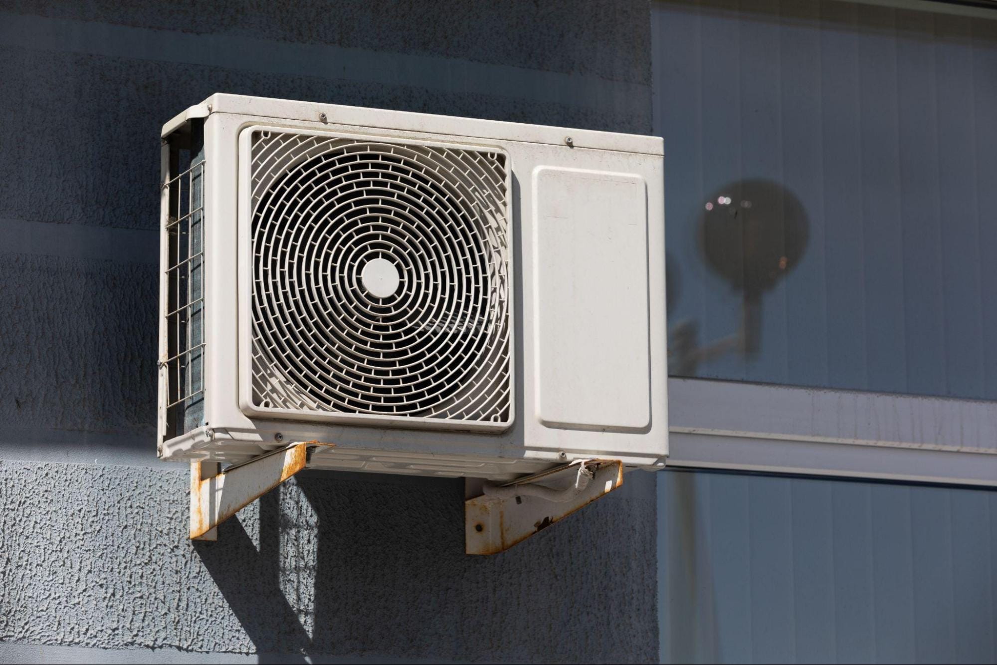 Close-up of an outdoor heat pump condenser fan mounted on rusted wall brackets against a home's exterior